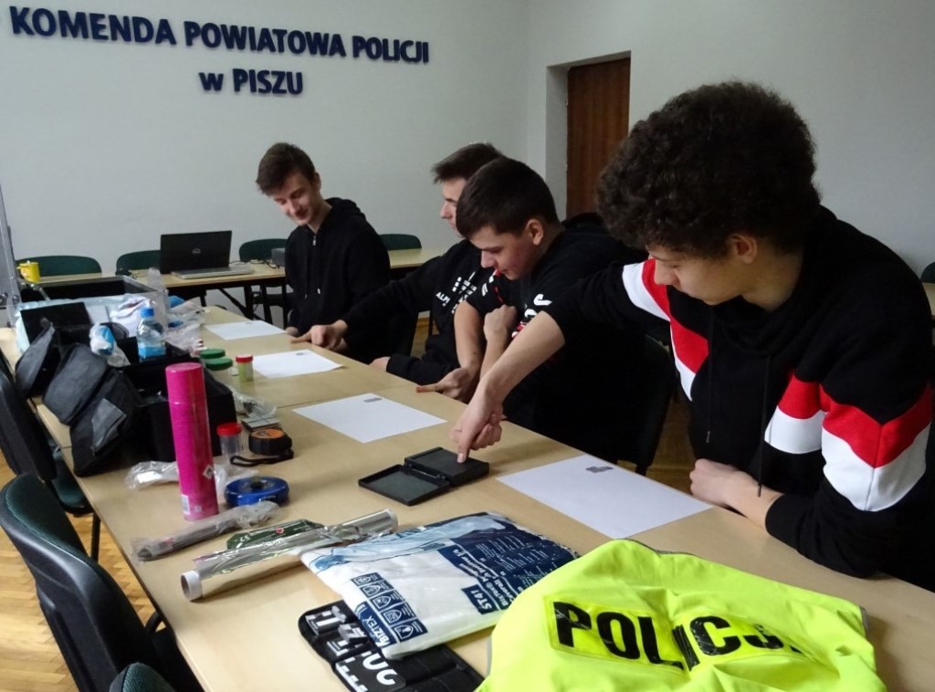 Dzień przedsiębiorczości - spotkanie z technikiem kryminalistyki - 4 uczniów siedzących przy stole odciska na białych kartkach swoje odciski palców, na stole leżą także różne przedmioty wykorzystywane w pracy technika oraz zielona kamizelka z napisem Policja