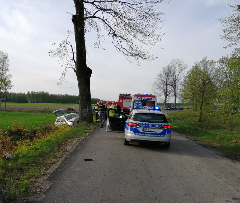Na pierwszym planie policyjny oznakowany radiowóz, na dalszym wóz  strażacki oraz strażacy, a po lewej stronie na poboczu drzewo, w które uderzyła osobowa skoda