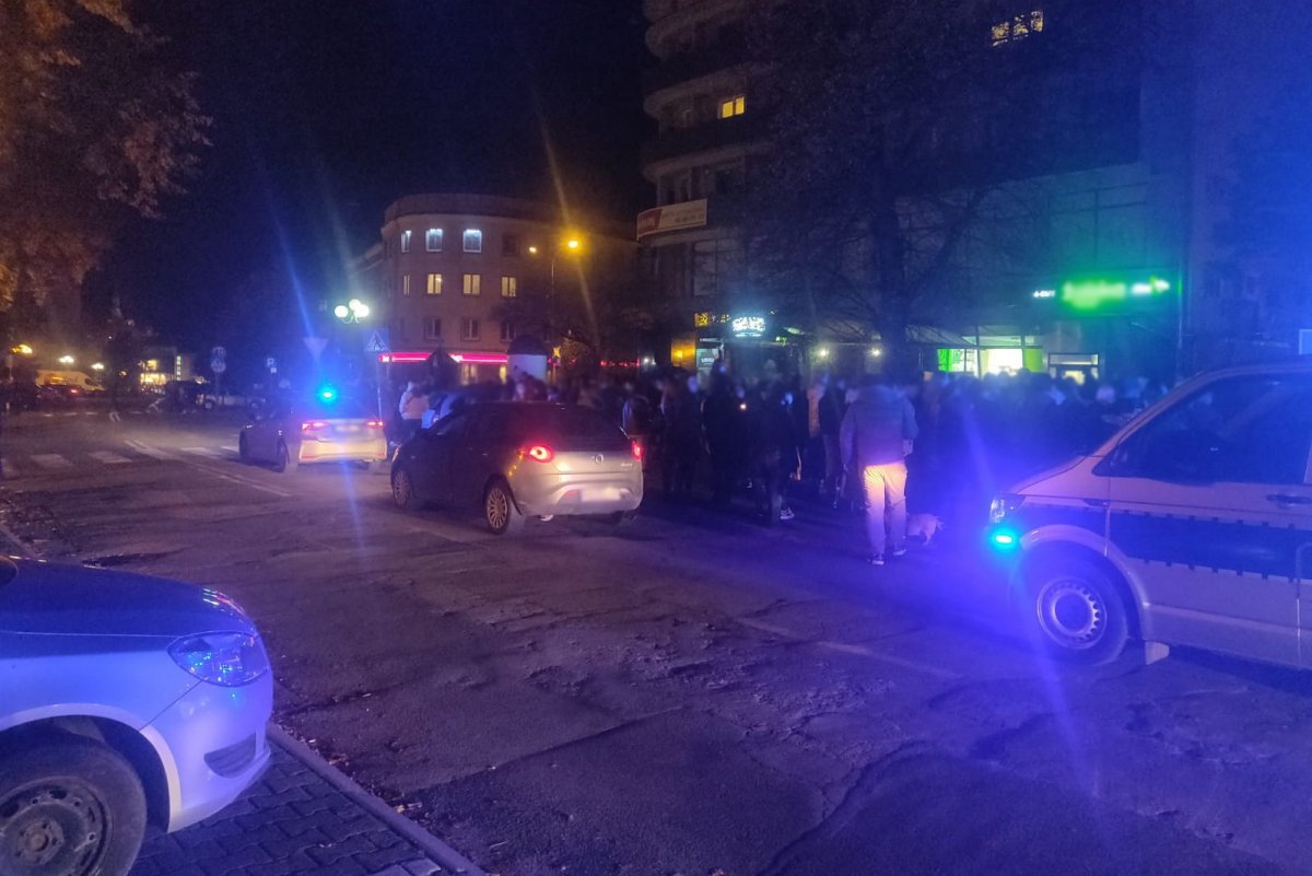 Policyjne radiowozy na miejscu nielegalnego zgromadzenia wieczorem na ulicy w Olsztynie, a za nimi uczestnicy zgromadzenia. Włączone sygnały świetlne radiowozów.