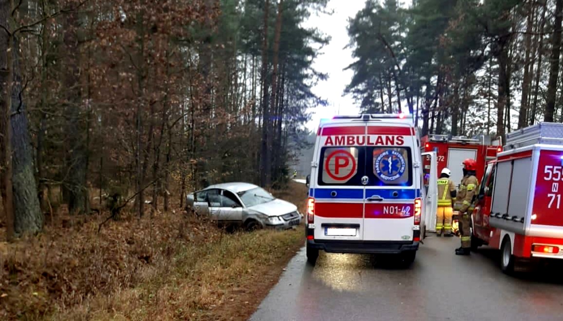 Na zdjęciu widać pojazd stojący w rowie po lewej stronie, po prawej stronie stoją dwa wozy strażackie i pojazd karetki pogotowia, pomiędzy pojazdami stoi 2 strażaków