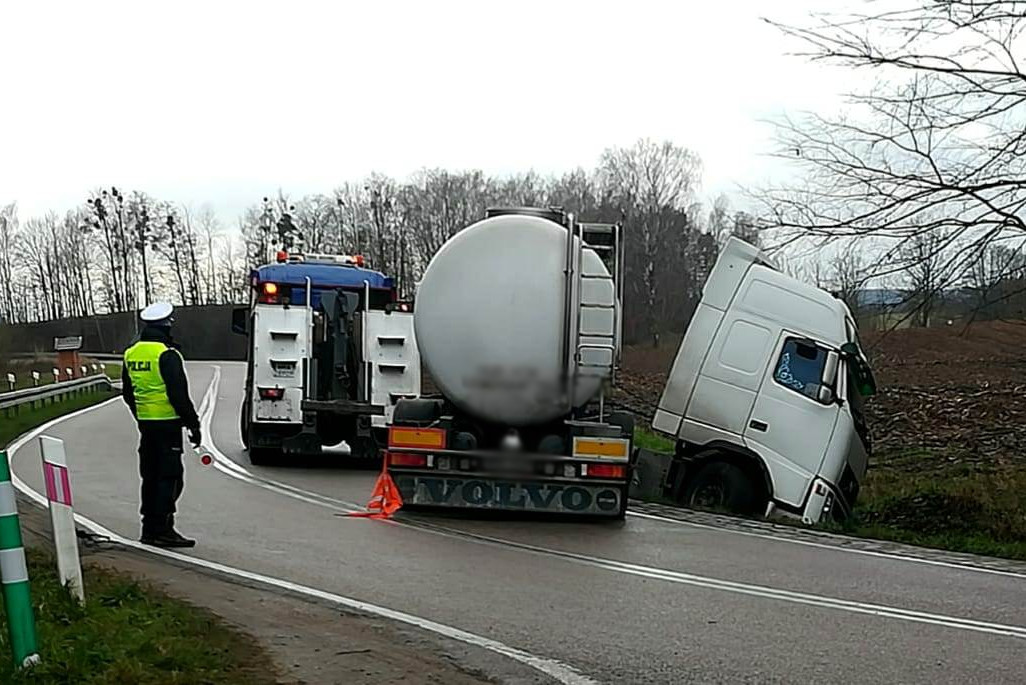 Miejsce zdarzenia drogowego w powiecie szczycieńskim. Ciężarówka częściowo stojąca na drodze, a częściowo w przydrożnym rowie a obok stojący policjant.