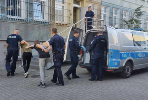 Zatrzymane osoby prowadzone przez policjantów z radiowozu do budynku komendy