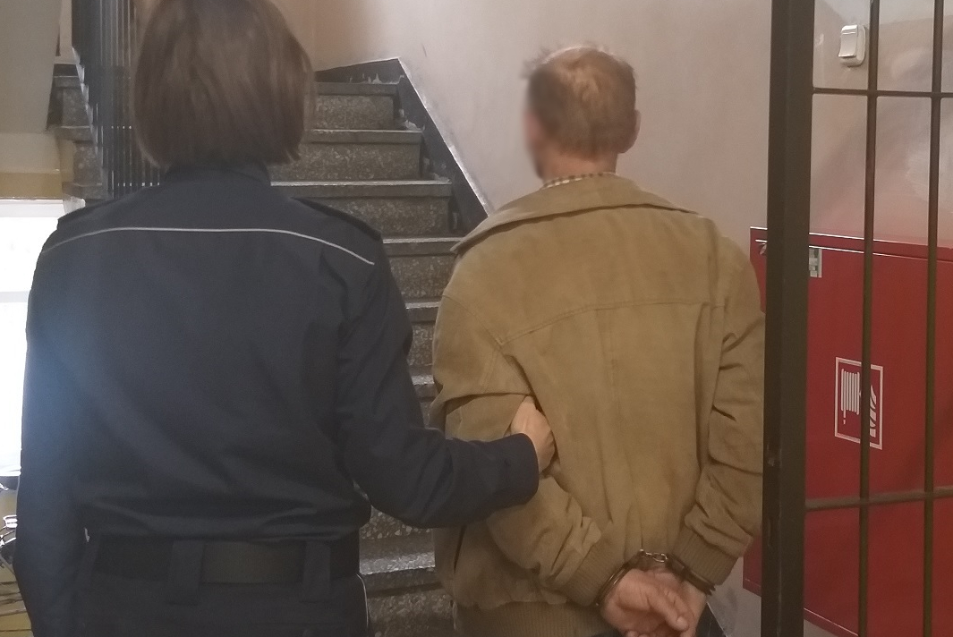 Mężczyzna zatrzymany za usiłowanie wręczenia łapówki policjantom, stoi na korytarzy trzymany przez policjantkę.