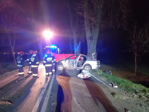 Rozbity samochód stojący na drodze oraz strażacy i wóz strażacki obok