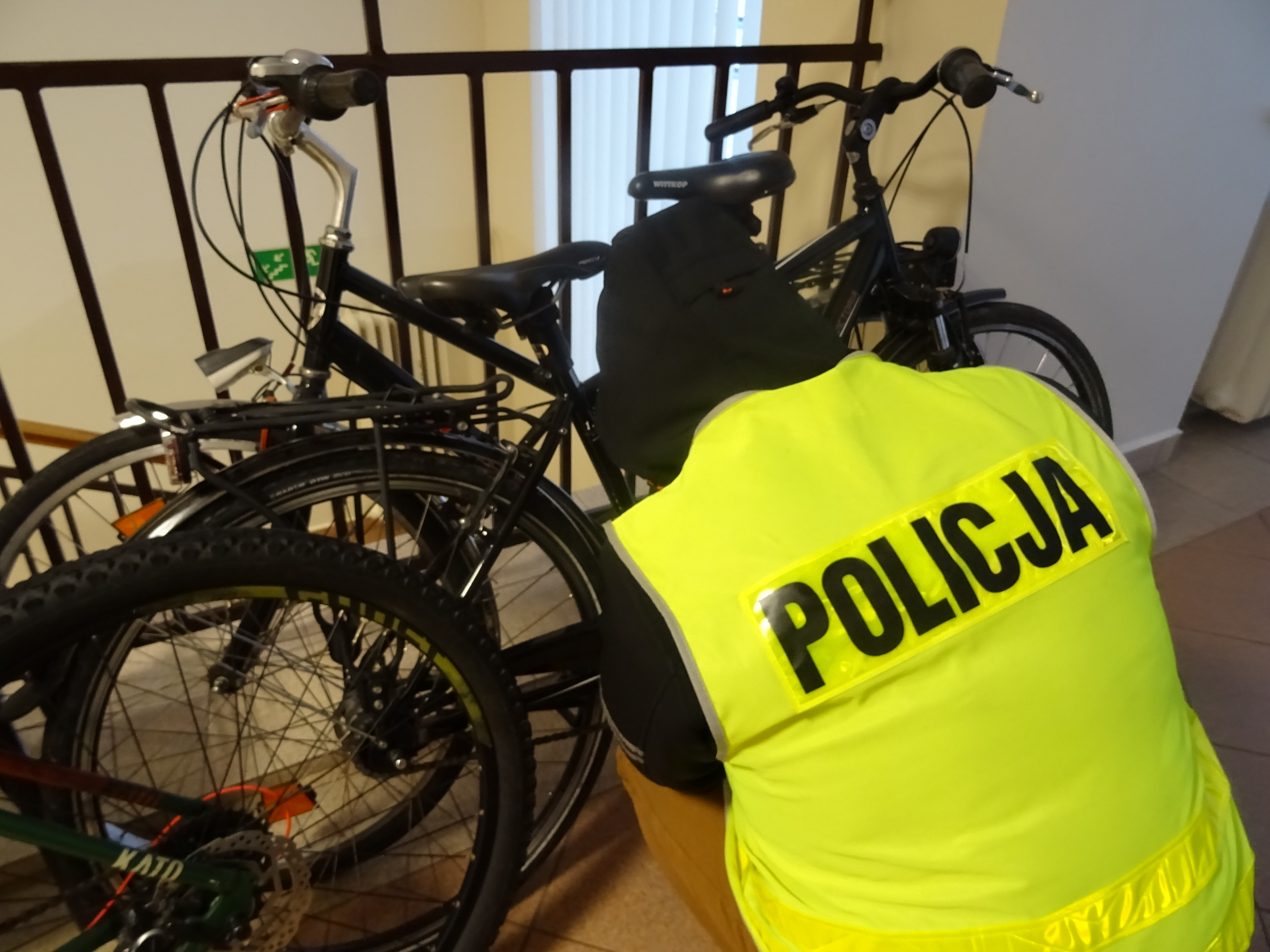 Oparte o barierkę dwa rowery koloru ciemnego i kucający przy nich policjant odwrócony tyłem w żółtej kamizelce z czarnym napisem Policja