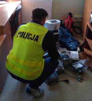 Policjant w żółtej kamizelce z napisem Policja kucający nad rozłożonymi na podłodze odzyskanymi przedmiotami pochodzącymi z kradzieży.