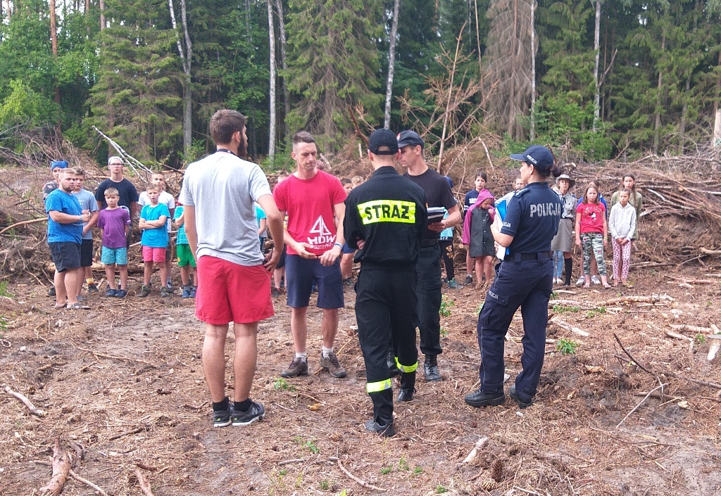 Policjantka, strażacy, pracownik sanepidu rozmawiają o bezpieczeństwie na obozie harcerskim w tle grupa kilkudziesięciu dzieci