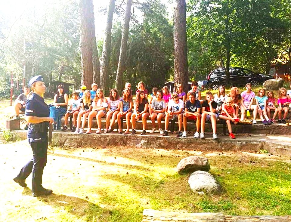 Policjantka stoi przed grupą młodzieży siedzącą na ławkach na podwórku i omawia zasady bezpiecznego wypoczynku nad wodą