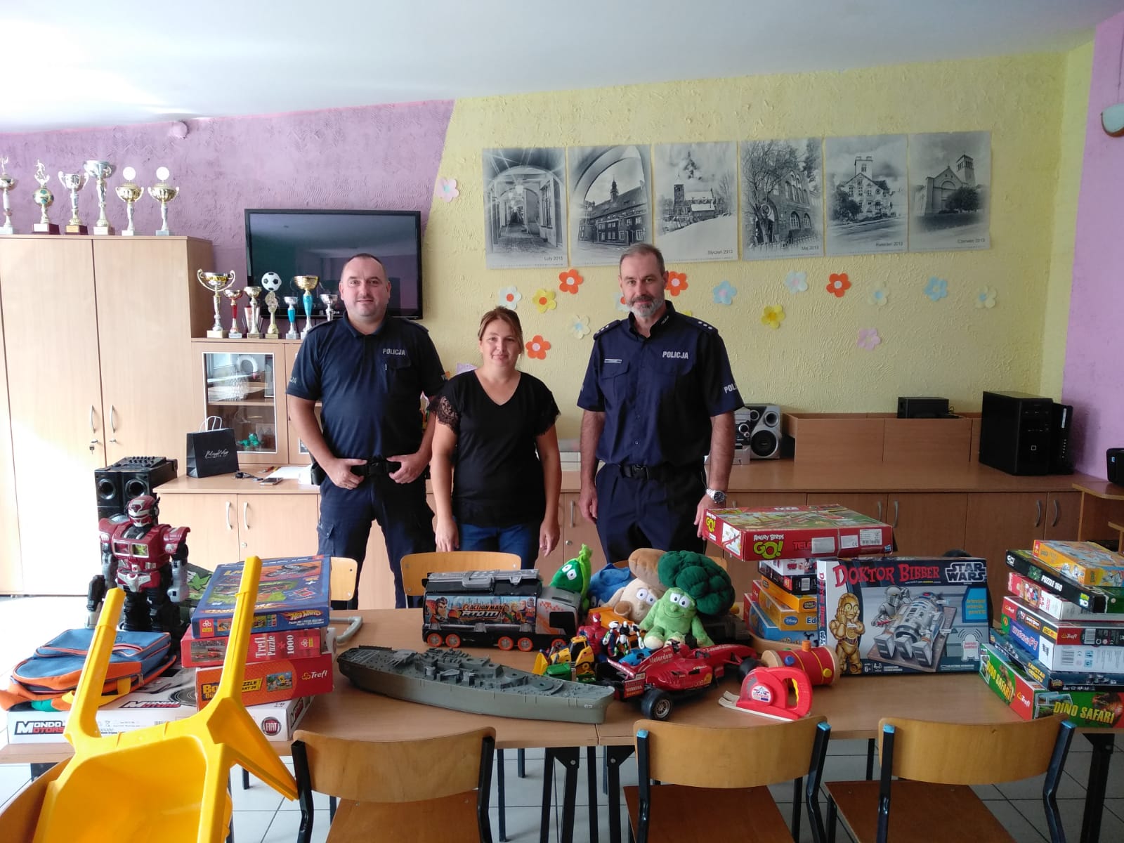 Trzy osoby, 2 policjantów i opiekunka świetlicy, a przed nimi rozłożone na stole zebrane, zabawki, puzzle, gry edukacyjne