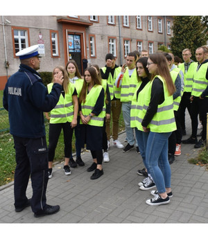Przed szkołą policjant rozmawia z uczniami ubranymi w kamizelki odblaskowe o bezpieczeństwie pieszych na drodze