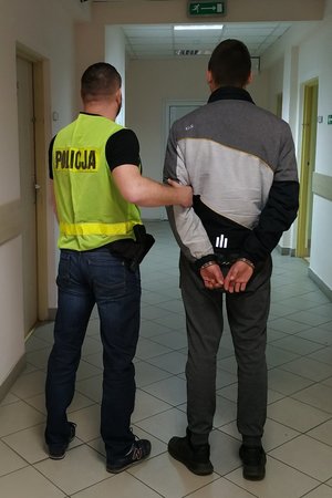 Zatrzymany 17-latek podejrzany o uszkodzenie mienia stoi tyłem, a za nim policjant operacyjny w kamizelce odblaskowej