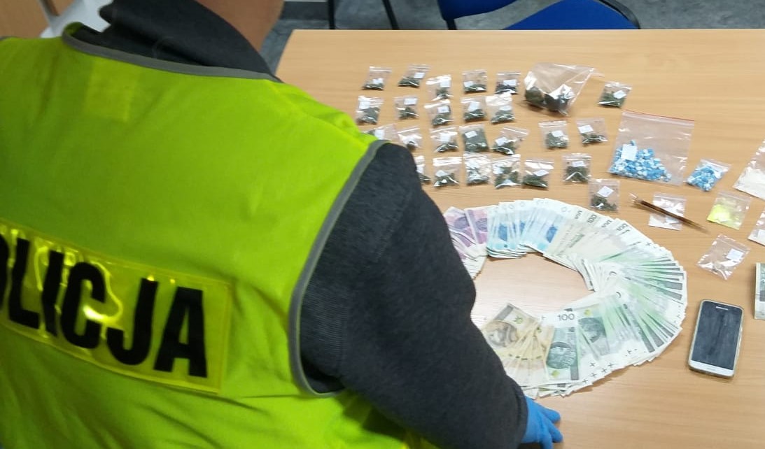 Na stole leżą rozłożone zabezpieczone banknoty i narkotyki, przy stole siedzi odwrócony policjant w żółtej kamizelce z napisem policja