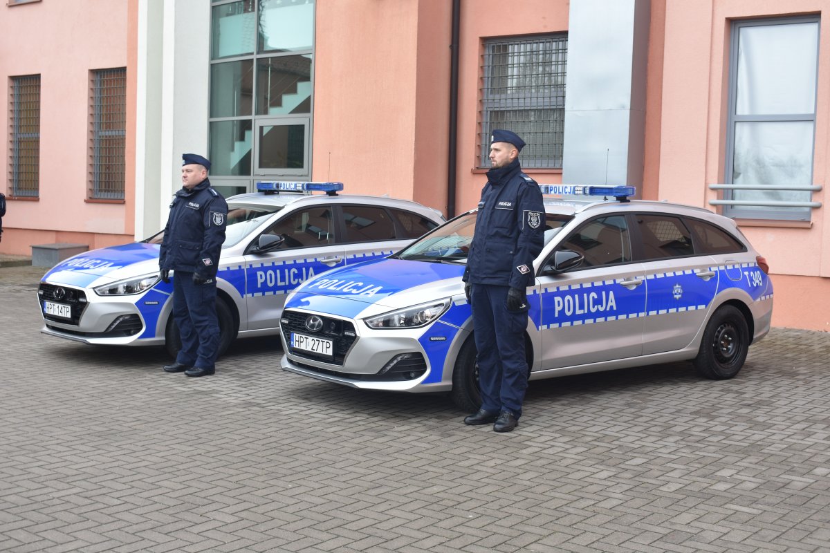 Dwa oznakowane radiowozy  marki Hundai i30 oraz przy każdym z nich stojący policjant