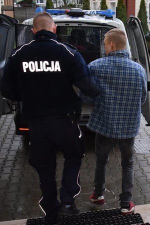 Przed wejściem do radiowozu stoi policjant z podejrzanym o groźby karalne