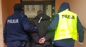 Dwóch policjantów  -jeden w granatowym umundurowaniu , drugi po cywilnemu w żółtej kamizelce z napisem policja - prowadzi do budynku mężczyznę podejrzanego o posiadanie narkotyków