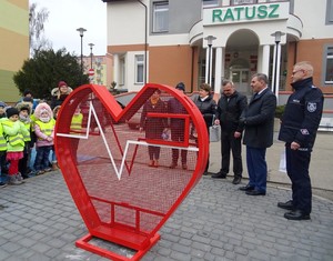 Metalowe serce na nakrętki, w tle stoi Komendant Powiatowy Policji w Piszu oraz Burmistrz Miasta Orzysz