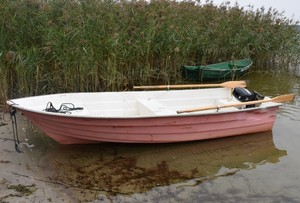 Na pierwszym planie na tafli jeziora łódka z dwoma wiosłami, w tle trzciny i druga łódka koloru zielonego.
