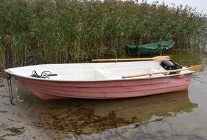 Na pierwszym planie na tafli jeziora łódka z dwoma wiosłami, w tle trzciny i druga łódka koloru zielonego.
