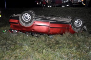 Nie pierwszym planie leży samochód w rowie kołami do góry, w tle stoi radiowóz strażacki i sześć osób.