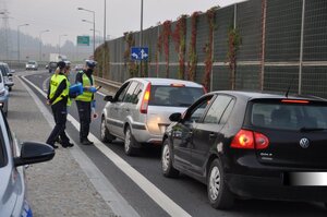 Zdjęcie przedstawia samochody osobowe stojące wzdłuż pasa drogowego, przy którym stoi dwóch funkcjonariuszy z urządzeniem do badania stanu trzeźwości.