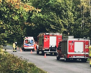 Na zdjęciu trzy pojazdy stojące na drodze. Jeden pojazd karetki pogotowia i dwa pojazdy straży pożarnej. Na środku drogi stoi dwóch strażaków.