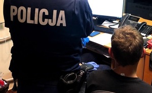 Na zdjęciu policjant w umundurowaniu stojący tyłem obok niego siedzi tyłem mężczyzna