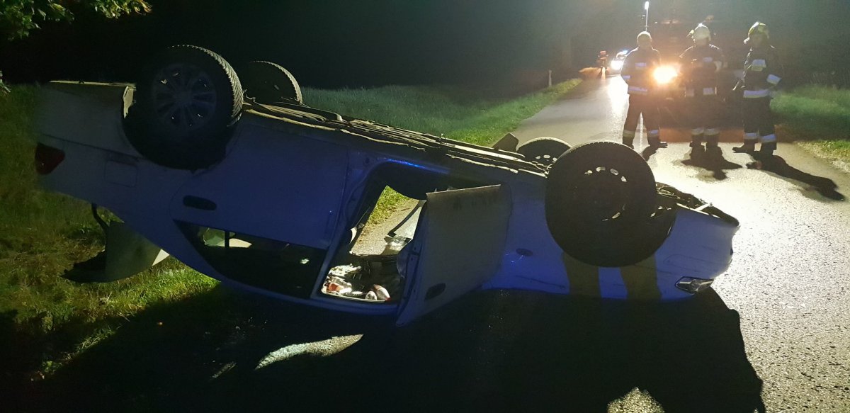 Na zdjęciu widać pojazd leżący na drodze kołami do góry, w tle stoi trzech strażaków