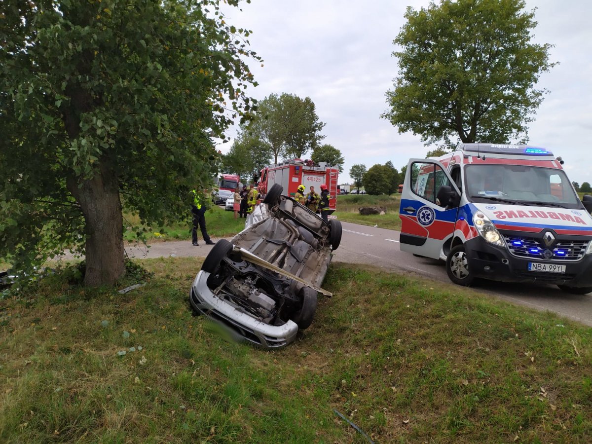 Na zdjęciu widać pojazd wywrócony kołami do góry leżący w rowie, w tle pojazd karetki pogotowia i straży pożarnej