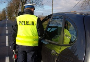 Przy pojeździe stoi policjant umundurowany tyłem w żółtej kamizelce z napisem policja