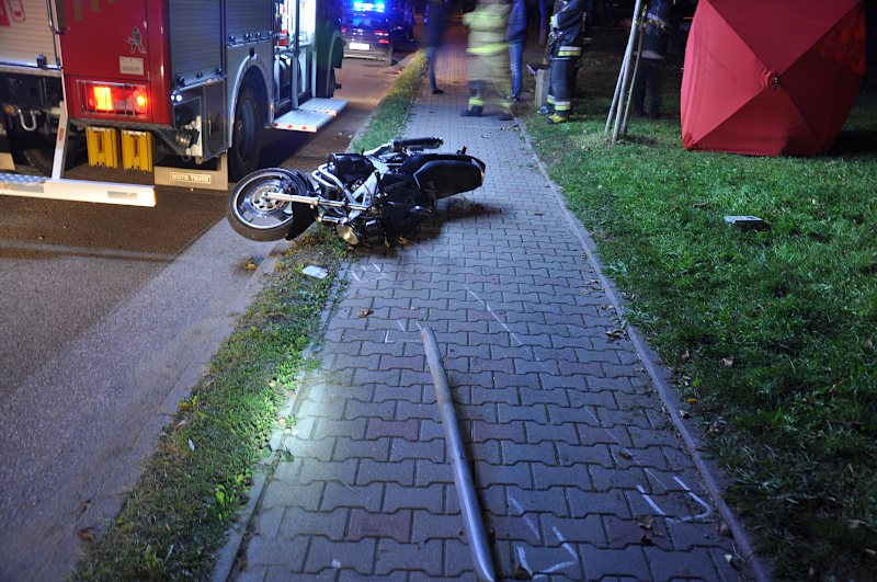 Na zdjęciu widać przewrócony motocykl, obok stoi wóz strażacki, w tle stoją osoby