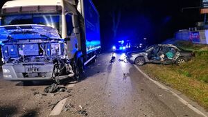 Na zdjęciu widać rozbity przód pojazdu ciężarowego i rozbity samochód osobowy leżący w rowie, w tle widać stojącego policjanta i radiowóz policyjny na sygnałach