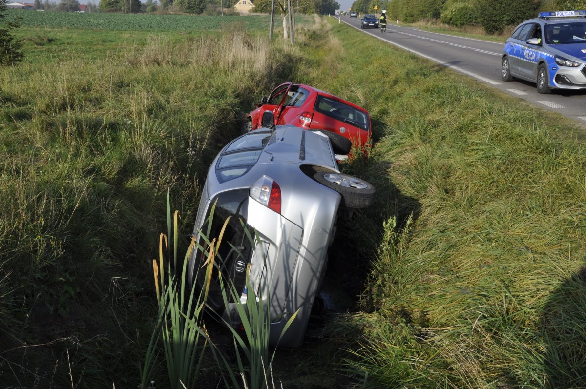 w rowie po lewej stronie drogi, na lewym boku leży srebrna honda, dalej rozbity czerwony VW, po prawej policyjny radiowóz, dalej na drodze stoi umundurowany strażak kierujący ruchem, przed nim zatrzymane czarne auto osobowe