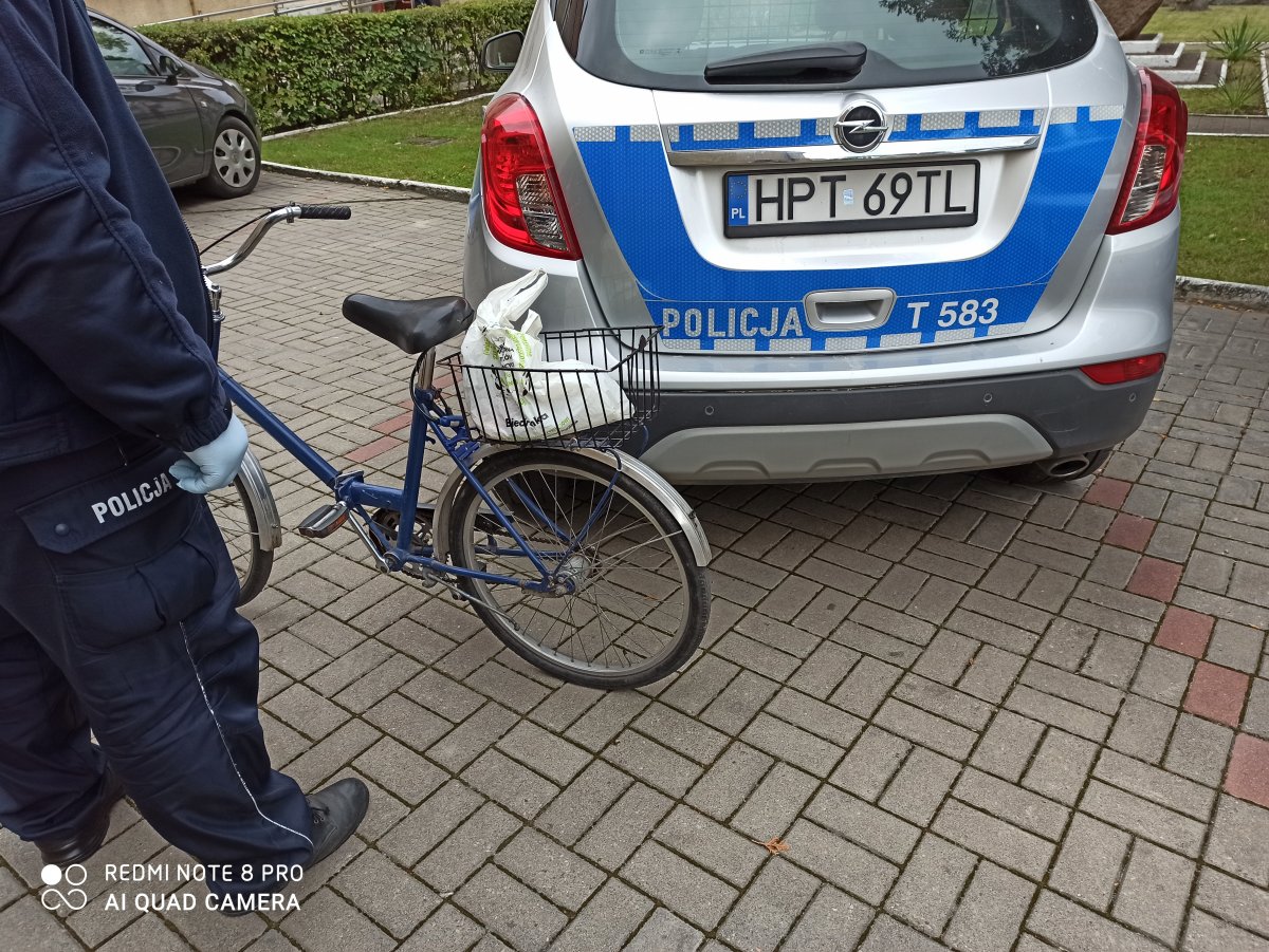 na zdjęciu policjant trzyma rower w kolorze niebieskim, obok stoi radiowóz