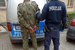 Na zdjęciu policjant trzyma pod rękę mężczyznę, przed nimi stoi oznakowany radiowóz