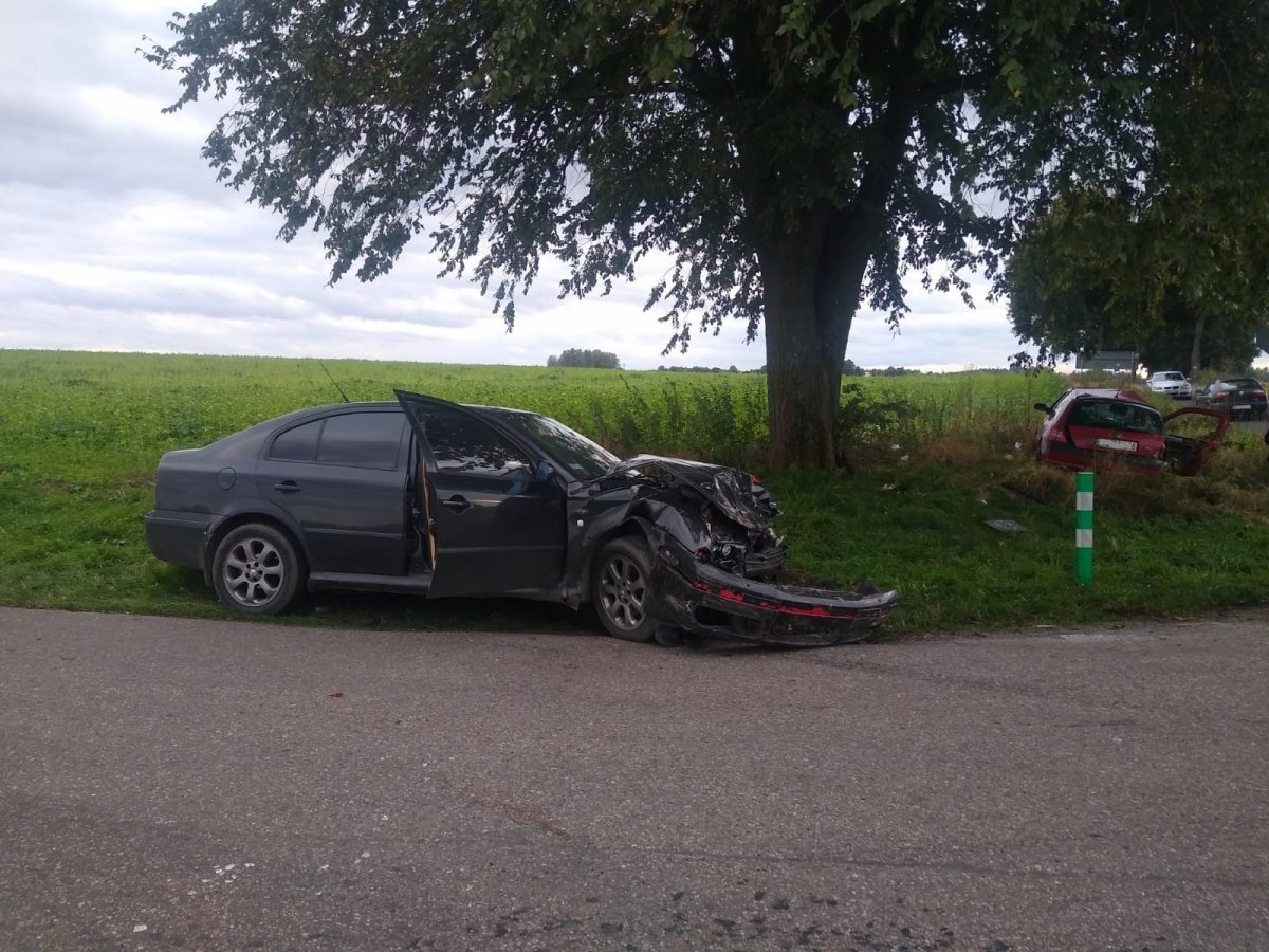 Na zdjęciu widać uszkodzony pojazd w kolorze czarnym, w tle stoi czerwony pojazd również uszkodzony podczas zdarzenia drogowego