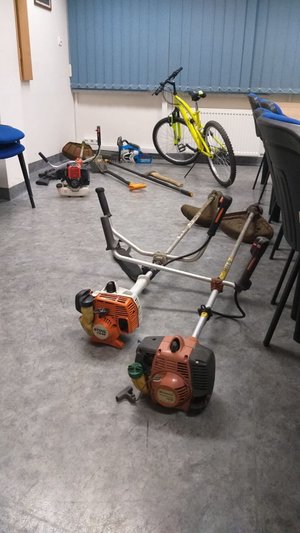 Na zdjęciu znajdują się rower górski, piła spalinowa, 3 podkaszarki spalinowe, wiatrówka i różnego rodzaju narzędzia