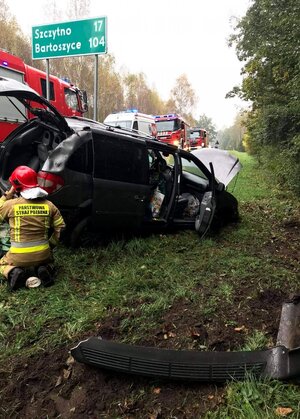 Samochód leżący w rowie, przy nim kuca strażak w tle stoją wozy strażackie