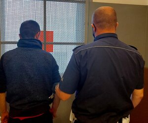 na zdjęciu widać dwóch mężczyzn stojących tyłem. policjant stojący po prawej stronie trzyma za rękę mężczyznę stojącego z lewej strony