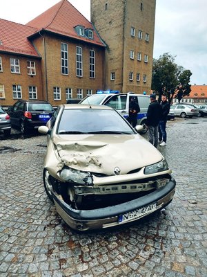 Na pierwszym planie stoi uszkodzony pojazd w tle, stoi budynek, radiowóz i zaparkowane pojazdy