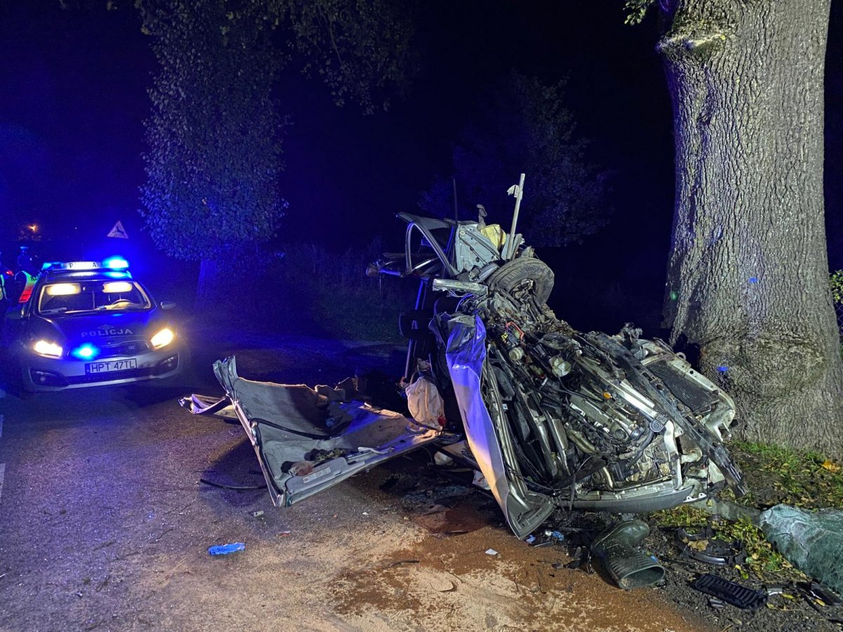 Na zdjęciu widać rozbite auto, po prawej stronie stoi drzewo po lewej radiowóz na sygnałach świetlnych