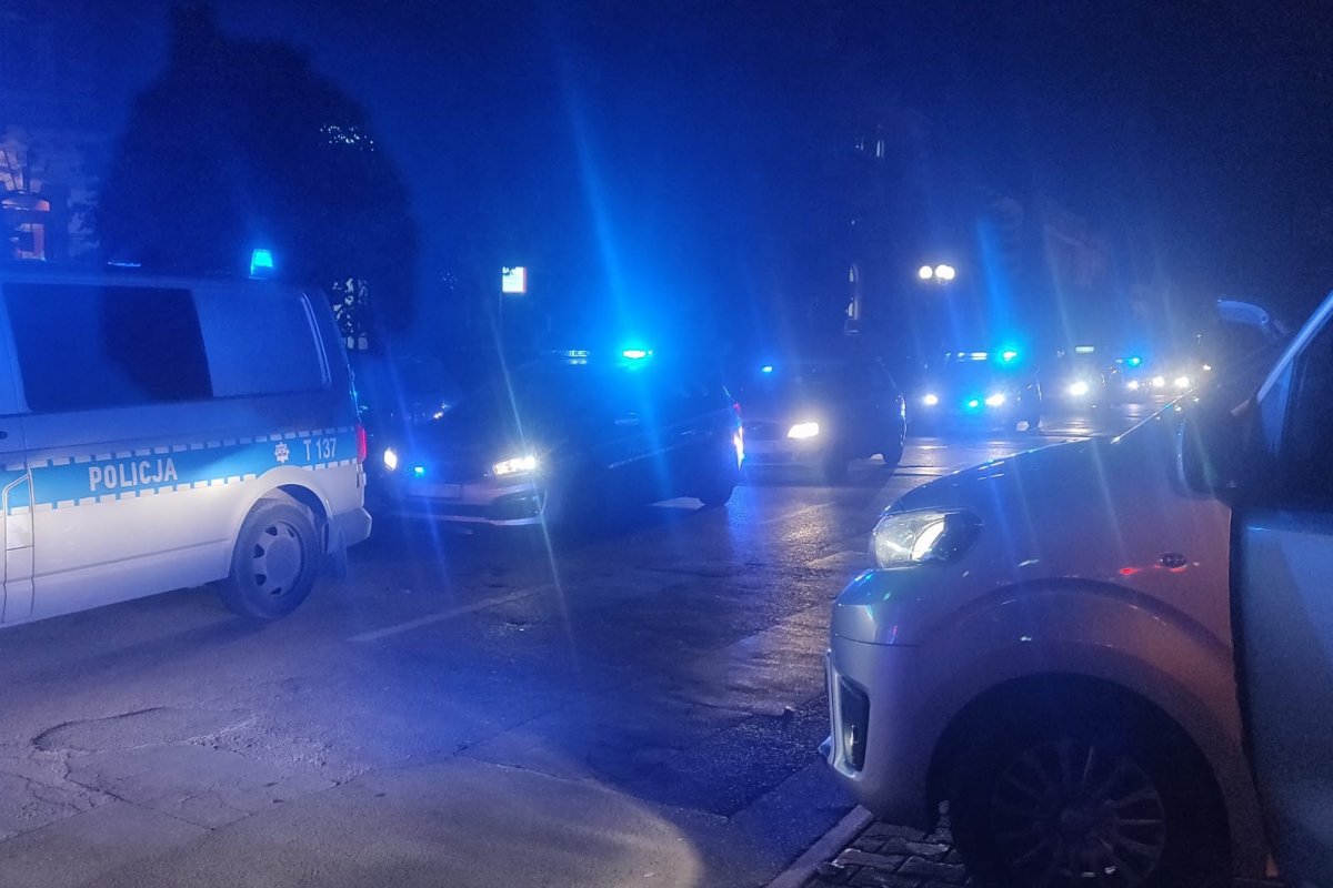 Policyjne radiowozy na miejscu nielegalnego zgromadzenia wieczorem na ulicy w Olsztynie, a za nimi uczestnicy zgromadzenia. Włączone sygnały świetlne radiowozów.