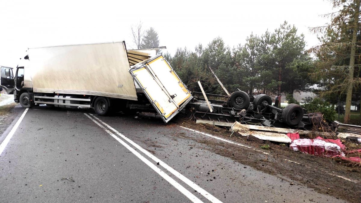 Na zdjęciu widać stojący na drodze uszkodzony pojazd ciężarowy i przewrócona naczepa, obok leżą połamane drzewa
