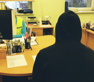 Mężczyzna siedzący przy biurku na głowie kaptur, przed nim siedząca policjantka przed komputerem