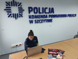 Na zdjęciu policjantka siedząca przed laptopem, w tle logo komendy powiatowej policji w Szczytnie