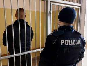 Mężczyzna stojący tyłem w celi, przed kratą stoi tyłem umundurowany policjant