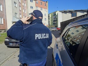 Policjant stojący tyłem przy samochodzie z ręką uniesioną z telefonem przy uchu, przed blokiem
