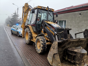 żółta koparka, za nią stoi radiowóz na sygnałach  świetlnych