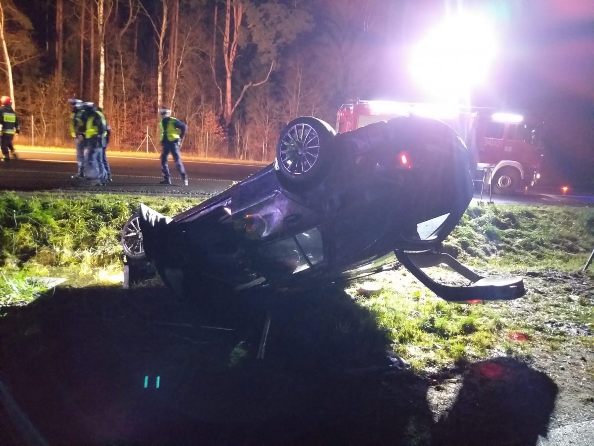 Na zdjęciu auto leżące na dachu, w tle służby ratunkowe