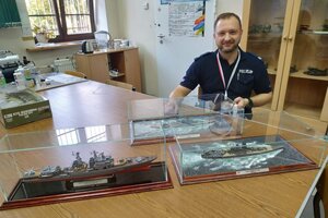 Sierż. szt. Rafał Szymborski siedzący za stołem na którym umieszczone są wykonane przez niego modele łodzi