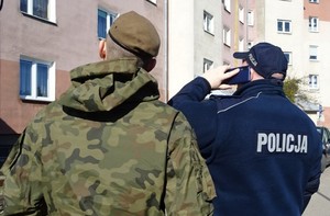Policjant stojący tyłem i trzyma telefon przy uchy, po lewej jego stronie stoi żołnierz WOT, przed nimi blok
